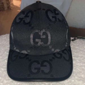 Svart keps från Gucci med GG-mönster - Snygg svart keps från Gucci med det klassiska GG-mönstret i grått och svart. Kepsen har böjd skärm, justerbar rem bak och är tillverkad i ett slitstarkt material. Perfekt för dig som vill ha en stilren accessoar med lyxig känsla.