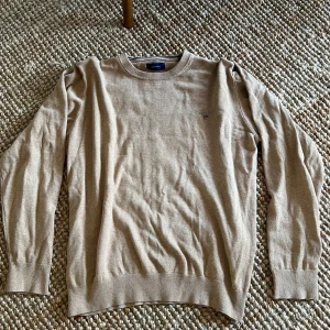 stickad tröja - Säljer en klassisk beige stickad tröja från GANT i storlek M. Tröjan har rund halsringning, ribbade muddar och en liten broderad logga på bröstet. Perfekt för en stilren och avslappnad look.🚀