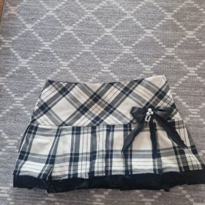 Rutig kortkjol med rosett och pärldetalj - Supersöt kortkjol i svart och vitt rutigt mönster. Kjolen har en dekorativ svart rosett med pärldetalj framtill och ett lager av svart spets längst ner. Perfekt för dig som gillar en söt och trendig stil. Dragkedja på sidan. Från h&m men är omsydd av mig. Tvätta försiktigt 