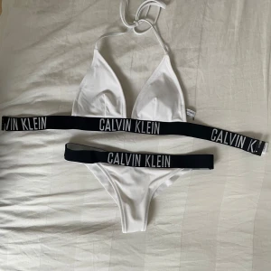 Vit bikini från Calvin Klein - Snygg vit bikini från Calvin Klein med svarta resårband där loggan är tryckt i vitt. Överdelen har trekantsform och knyts i nacken. Perfekt för sommaren och strandhäng.