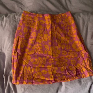 Rosa och gul blommig kjol från Bik Bok - Säljer en kort kjol från Bik Bok i rosa tyg med stora gula blommor. Kjolen har en liten slits framtill och dragkedja i sidan. Perfekt för dig som gillar färgglada och unika plagg.