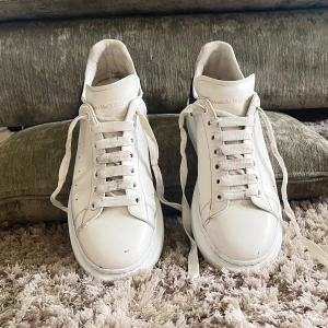 Vita sneakers från Alexander McQueen - Säljer ett par klassiska vita sneakers från Alexander McQueen med svart hälparti och guldfärgad logga. Skorna har rund tå, platt sula och snörning. Perfekta för en stilren och trendig look. Skriv privat om priset och om du vill ha fler bilder