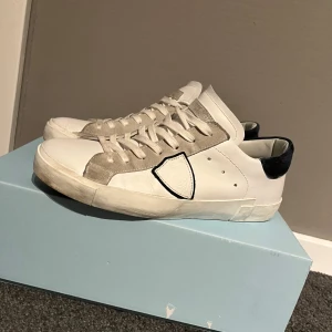 Vita sneakers från Philippe Model - Säljer ett par vita Philippe Model sneakers med grå mockadetaljer och svart häl. Skorna har snörning, rund tå och platt sula skorna är i nytt skick och säljs pga för små skorna kostar 3000kr nya 