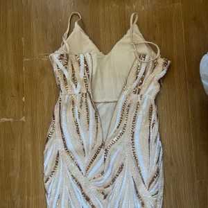 Vit långklänning med paljetter - Säljer en elegant vit långklänning med smala axelband och djup v-ringning. Klänningen har ett mönster av vita och guldiga paljetter som ger en glittrig effekt. Perfekt för bal eller fest. Figurnära passform och slits framtill för extra stil.