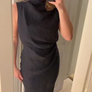Svart omlottklänning med drapering - Säljer en svart klänning med hög hals och snygg drapering framtill. Klänningen har ärmlös design och omlott-detalj i midjan. Materialet har en lätt glansig yta och subtil struktur som ger en lyxig känsla. Helt ny med prislapp, skicka för fler bilder 