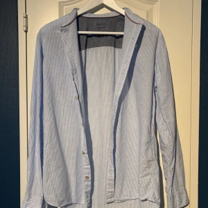 Blårandig skjorta från Essentials - Säljer en blårandig skjorta från Essentials med bandkrage och knappar framtill. Skjortan har tunna vita och blå ränder. Perfekt för en stilren look. 