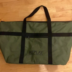Olivgrön weekendbag från Replay - Stor olivgrön weekendbag från Replay med svarta handtag och detaljer. Väskan har en dragkedja upptill, en ficka med dragkedja framtill och en stor ytterficka på baksidan. Perfekt för resor eller gymmet.