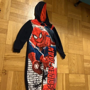 Spiderman hoodie jumpsuit 134cm - Supercool Spiderman hoodie jumpsuit i marinblått med stora röda och blå detaljer samt tryck av Spiderman på framsidan. Långärmad med huva som har röd insida. Mjuk och skön, perfekt för chilliga dagar eller som pyjamas.