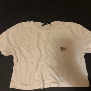 Vit croppad t-shirt Brandy Melville - Vit croppad t-shirt från Brandy Melville med liten broderad amerikansk flagga på bröstet. Klassisk rund hals och korta ärmar. Tillverkad i 100% mjuk bomull, perfekt för chill dagar. Enkel och clean design som funkar till jeans eller shorts.
