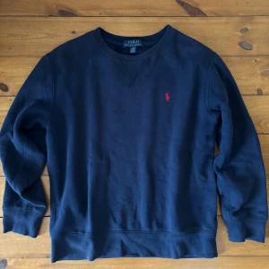 Mörkblå sweatshirt från Polo Ralph Lauren - Klassisk mörkblå sweatshirt från Polo Ralph Lauren med rund halsringning och röd broderad logga på bröstet. Tröjan har långa ärmar och ribbade muddar vid ärmslut och nederkant. Perfekt för en chill och stilren look.
