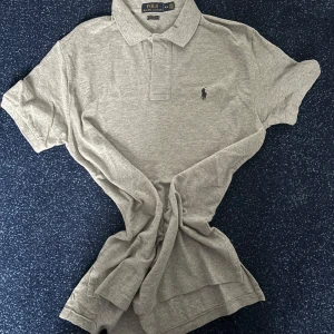 Grå pikétröja från Polo Ralph Lauren - Klassisk grå pikétröja från Polo Ralph Lauren i bomull. Tröjan har korta ärmar, krage och knäppning framtill samt den ikoniska broderade loggan på bröstet. Classic fit för en avslappnad stil.