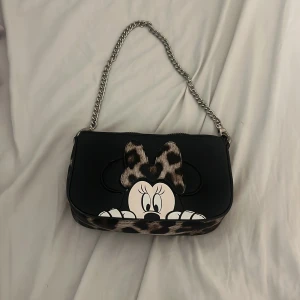 Disney axelväska med leoparddetaljer - Snygg svart axelväska från Disney med kedjerem och coolt tryck av Minnie Mouse. Bågen och botten har leopardmönster och väskan har både en kedja och en bredare axelrem. Matchande rund plånbok medföljer, också med Minnie och leoparddetaljer.