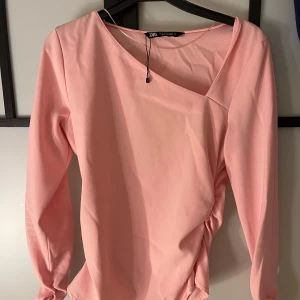 Ljusrosa omlottopp från Zara - Trendig ljusrosa omlottopp från Zara med lång ärm och snygg asymmetrisk halsringning. Toppen har en lätt rynkad detalj i sidan som ger en cool twist. Perfekt för dig som gillar stilrena men unika plagg.