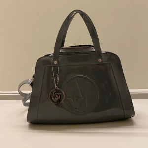 Grå handväska från Armani Jeans - Snygg grå handväska från Armani Jeans  med präglad logga framtill och matchande hänge. Väskan har två handtag, dragkedja upptill och ett rymligt foder med AJ-mönster samt innerfack med dragkedja. Perfekt för dig som gillar stilrena accessoarer. Priset är ej hugget i sten. Hör av er vid frågor eller funderingar! 