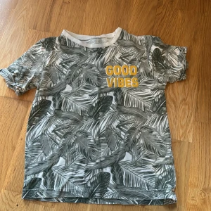 T-shirt med palmblad och text - Snygg t-shirt med grått palmbladsmönster på vit botten och gul text 'GOOD VIBES' på bröstet. Klassisk passform med korta ärmar och rund halsringning. Perfekt för dig som gillar en chill och tropisk stil.