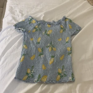 Blå smockad topp med citronmönster - Supersöt ljusblå topp med smockad struktur och korta ärmar. Toppen har ett lekfullt citronmönster i gult och grönt samt vågiga kanter vid hals, ärmar och nederkant. Perfekt för dig som vill sticka ut med en färgglad och somrig vibe.