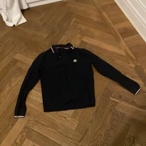 Svart Moncler pikétröja med röd/vit detalj - Snygg svart pikétröja från Moncler med långa ärmar. Kragen har röda och vita ränder som ger en sportig vibe. Tröjan har knappar framtill och Moncler-logga på bröstet. Perfekt för dig som gillar stilrena märkeskläder.