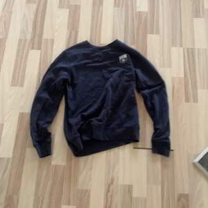 Mörkblå sweatshirt från GANT - Snygg mörkblå sweatshirt från GANT med rund hals och ett litet GANT-märke på bröstet. Tröjan har långa ärmar med quiltade detaljer på armbågarna och ribbade muddar. Perfekt för dig som gillar klassisk och stilren design.