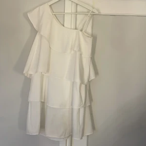 Vit volangklänning från Gina Tricot - Säljer en vit kortklänning från Gina Tricot med ”one-shoulder” och volangdetaljer. Klänningen har ett axelbandslöst snitt på ena sidan och ett smalt axelband på andra, vilket ger en snygg asymmetrisk look. Materialet är mjukt och lätt, perfekt för varma dagar. Gör dej redo redan nu för kommande fester/avslutningar. Sparsamt använd. 