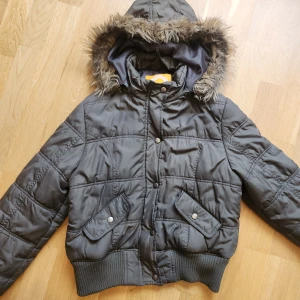 Snygg pufferjacka med fuskpäls - Säljer en mörkgrön pufferjacka från UPS! by UPS! med huva och fuskpäls. Jackan har ribbade muddar, två fickor med lock och knappar framtill. Perfekt för kalla dagar och har en snygg, lite kortare passform. Köpt på vinted för ett år sen. Passar S/M. 