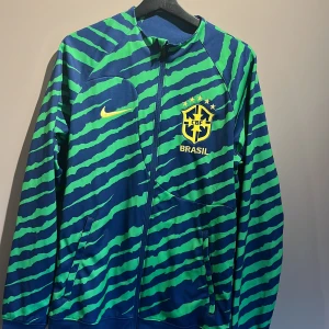 Brasil långärmad fotbollströja Nike S - Slim fit Brasil fotbollströja från Nike med dragkedja, lång ärm och coolt randigt mönster i marinblått och grönt. Broderat Brasil-märke med fem stjärnor och gul Nike-logga på bröstet. Tillverkad i polyester, perfekt för dig som gillar unik stil. Helt oanvänd köpt på sports direct i London storlek S