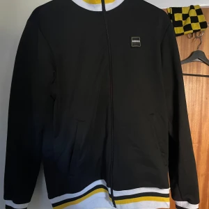 Svart Norra Staplats hoodie med dragkedja - Svart hoodie från Norra Staplats med hel dragkedja och ribbade muddar i vitt, gult och svart. Märke på bröstet och två fickor framtill. Snygg streetstil med långärmad passform och coola kontrastdetaljer. Perfekt för dig som gillar sportiga och unika plagg.