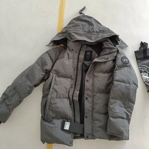 Grå dunjacka XXL - Säljer en grå dunjacka i storlek XXL. Jackan har huva, dragkedja och knappar framtill samt klassisk logga på ärmen. Perfekt för kalla dagar och har en stilren look med pösig passform.