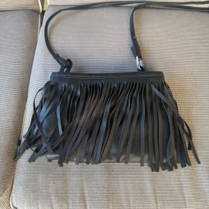 Svart axelväska med fransar - Snygg svart axelväska i skinn med långa fransar framtill som ger en cool och trendig look. Perfekt storlek för det viktigaste och med justerbar axelrem. Väskan har en stilren design som sticker ut med sina fransdetaljer.