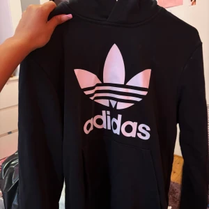Svart Adidas hoodie med stor logga - Svart hoodie från Adidas med stor vit Trefoil-logga och text på bröstet. Klassisk känguruficka framtill och huva. Perfekt för dig som gillar streetwear och vill ha en enkel men snygg tröja. Mjuk och skön bomullskvalitet.