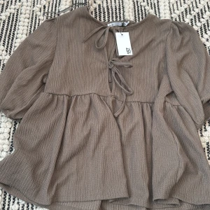 Brun blus med knyt från 157, S - Supersöt brun blus från 157 med volang nedtill och trendiga knytband framtill. Blusen har lång ärm och är tillverkad i ett ribbat material som ger en snygg struktur. Perfekt för dig som gillar en avslappnad men ändå stilsäker look.