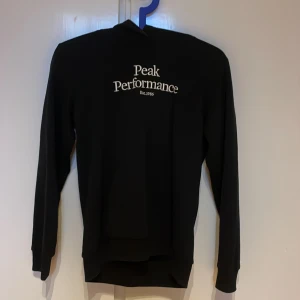 Svart hoodie från Peak Performance - Svart hoodie från Peak Performance med vit logga på bröstet. Passar även S! Klassisk modell med huva och långa ärmar, perfekt för chill dagar. Kom dm vid minsta fundering! Mjuk och skön känsla, passar dig som gillar enkel och stilren streetwear. Nypris 799!