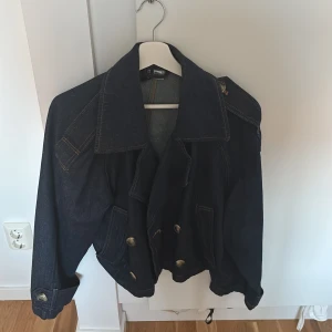 Oversized jeansjacka med stora knappar - Snygg oversized jeansjacka i mörkblå denim med stora guldiga knappar framtill och på ärmsluten. Jackan har bred krage och markerade sömmar som ger en cool vintagekänsla. Perfekt för dig som vill ha en statementjacka till din outfit.