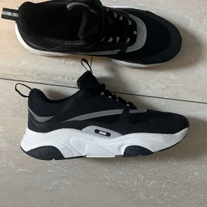 Svarta D sneakers med vit sula - Chunky svarta D sneakers med vit och svart sula, reflekterande detaljer och meshpaneler. Skorna har snörning och en sportig vibe med tydlig D-logga på sidan och CD-märkning undertill. Perfekta för dig som gillar exklusiva och trendiga sneakers.