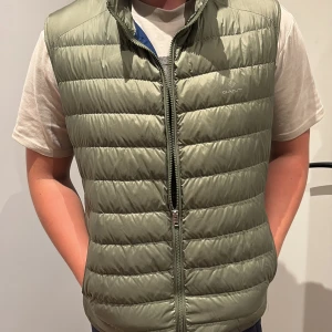Gant väst - Snygg olivgrön dunväst från GANT med quiltad design och dragkedja framtill. Västen har två fickor på sidorna och en diskret logga på bröstet. Insidan är fodrad i marinblått tyg. Perfekt för lager-på-lager och chill stil. Ny pris runt 2000 kr pris kan diskuteras vid snabb affär 😁