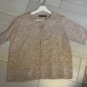 Beige kofta från 157 med pärlknappar - Mysig beige kofta från 157 i stickat material. Koftan har rund halsringning, ribbade muddar och fina pärlknappar framtill. Perfekt att slänga på sig när det är lite kyligt ute.