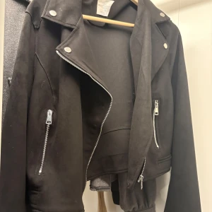 Svart bikerjacka i mocka-look - Snygg svart bikerjacka från Chiquelle i mocka-liknande material. Jackan har silvriga dragkedjor, klassisk krage med tryckknappar och croppad passform. Perfekt för dig som vill ha en edgy stil med stilrena detaljer. 