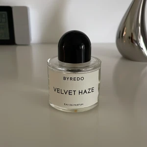 Byredo Velvet Haze Eau de Parfum 50ml - Byredo Velvet Haze Eau de Parfum i 50 ml flaska. Doften kommer i en stilren, rund glasflaska med svart kupolformad kork och vit etikett. Parfymen är tillverkad i Frankrike och har en transparent, ljusgul vätska. Perfekt för dig som gillar exklusiva och moderna dofter.