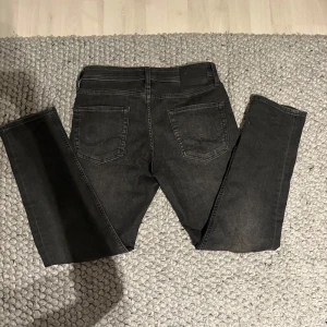 Svarta jeans från Jack & Jones - Säljer ett par tapered/mike svarta jeans från Jack & Jones i 30/30 med klassisk femficksdesign och smal passform. Jeansen har en mörk tvätt och raka ben, perfekt för dig som gillar en enkel och stilren look. Materialet är mjukt och stretchigt jeans för extra komfort.