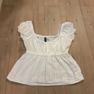 Vit blus med puffärmar från H&M - Supersöt vit blus från H&M med puffiga korta ärmar, smockad resår vid halsringningen och midjan samt knappar framtill. Tunt, luftigt bomullstyg med diskreta ränder och romantisk känsla. Perfekt för en somrig look!