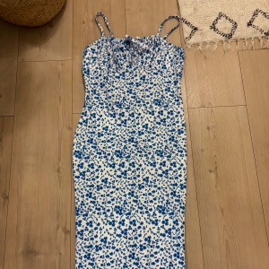 Vit och blå blommig långklänning - Supersöt långklänning med smala axelband och blått blommönster på vit botten. Klänningen har en figurnära passform och rynkdetalj vid bysten. Perfekt för varma dagar och somriga vibes.
