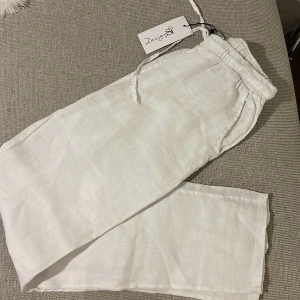 Vita linnebyxor wide från Design by Si - Snygga vita linnebyxor från Design by Si i modellen Classic Wide. Byxorna har resår och snörning i midjan för skön passform och är tillverkade i 100% linne. Perfekta för en chill och stilren look under varma dagar.