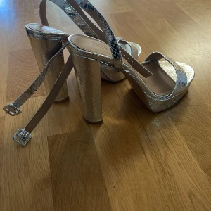 Silvriga platåsandaler med hög klack - Snygga sandaler i metallic silver med ormskinnsmönster och bred platåsula. Skorna har hög blockklack, öppen tå och smala remmar med spänne runt vristen. Perfekta för att sticka ut och ge extra höjd till din outfit.