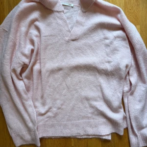 Ljusrosa stickad tröja från 157 - Supermjuk ljusrosa stickad tröja från 157 med v-ringning och krage. Tröjan har långa ärmar och ribbade muddar vid ärmslut och nederkant. Perfekt för dig som gillar en chill och snygg look.