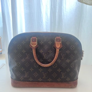 Louis Vuitton Monogram handväska - Klassisk handväska från Louis Vuitton i brunt skinn med ikoniskt monogrammönster och detaljer i ljusbrunt läder. Väskan har rundad form, dubbla handtag och guldig dragkedja med LV-logga. Insidan är rymlig med en innerficka för småsaker. Jag köpte den av en vän som köpte den i butik, men har inget kvitto tyvärr. Hör gärna av er om du är intresserad eller vill diskutera priset.