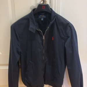 Mörkblå vindjacka från Polo Ralph Lauren - Snygg mörkblå vindjacka från Polo Ralph Lauren med klassisk röd broderad logga på bröstet. Jackan har dragkedja, två fickor och rutigt innerfoder. Perfekt för dig som gillar stilren och tidlös design.