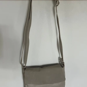 Grå axelväska i skinn - Snygg beige axelväska i  skinn (inte riktigt) med justerbar rem. Väskan har flera fack, både med och utan dragkedja, och en stilren design som passar till det mesta. Perfekt storlek för att få plats med allt du behöver till vardags. Använd endast en gång så är fortfarande i toppskick!🥰