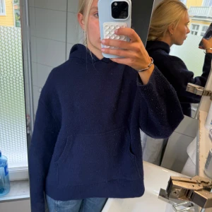 Mörkblå stickad hoodie med huva - Mysig mörkblå stickad hoodie med huva och stor känguruficka framtill. Tröjan har en loose passform och långa ärmar. Enkel och stilren design som funkar till jeans eller mjukisbyxor. Stoleken är S-M