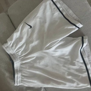 Vita mesh-shorts från Nike - Säljer ett par vita mesh-shorts från Nike med svart logga och svarta detaljer längs kanterna. Shortsen har elastisk midja och är tillverkade i lätt och ventilerande polyester, perfekta för sport eller chill. Klassisk loose fit och stilren design. Lite små fläckar