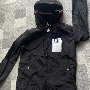 Svart vindjacka från Moncler med huva - Svart vindjacka från Moncler med huva och dragsko, klassisk logga på ärmen och snygg randig detalj i rött, vitt och blått på huvan. Jackan har dragkedja framtill, två fickor med tryckknappar och är tillverkad i lätt polyester. Perfekt för dig som gillar stilren och sportig look.