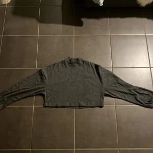 Grå croppad ribbad tröja FB Sister från New Yorker 60kr + frakt  - Mörkgrå croppad tröja från FB Sister i storlek XS. Tröjan är ribbstickad med lång ärm och har en högre hals. Perfekt för dig som gillar en chill och trendig stil. Materialet är mjukt och stretchigt, vilket gör den superbekväm.